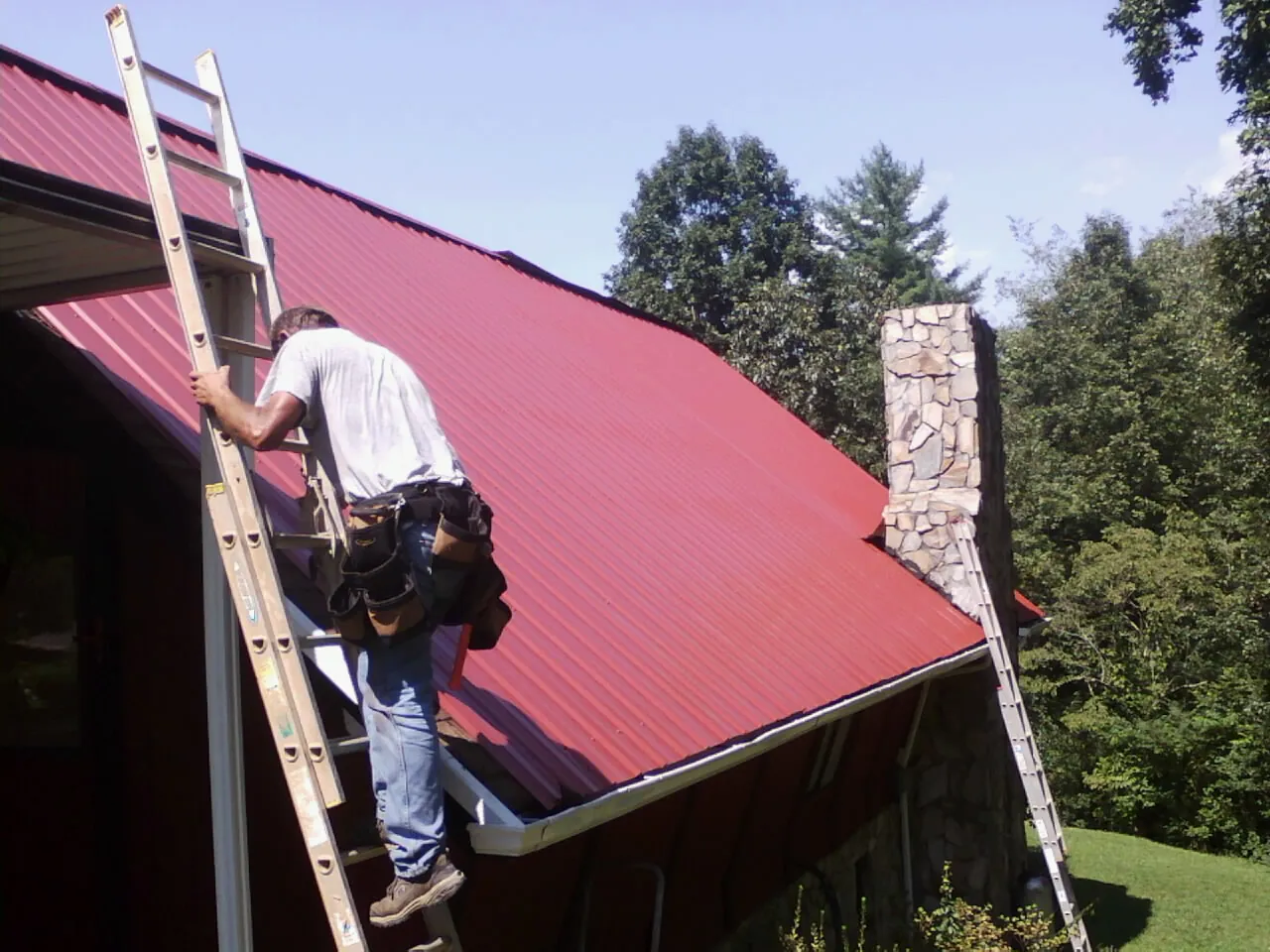 Roofer on a ladder beside a red metal roof and stone chimney — Soffit & Fascia Repair by Roofing Brothers in Smicksburg